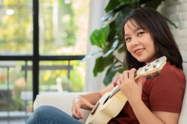 Mutlu Asyalı kadın oturma odasında kanepede oturuyor ve rahatça ukulele çalışıyor. Sanatçıdan rahatlama ve ilham. Müzik kavramı, rahatlama ve hobi aktiviteleri boş zamanların tadını çıkar