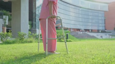 Yaşlı bir Asyalı kadın yürüteç kullanıyor ve hastanenin arka bahçesinde yürüyor. Fizik tedavi, rehabilitasyon ve sağlık sigortası konsepti. Yaşlı bakımı için tıbbi kemik iltihabı..