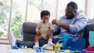 Mutlu Afrikalı Amerikalı adam oturma odasındaki kanepede küçük bebek bebekle oynuyor. Ebeveyn ve küçük çocuk evde dinleniyor. Ebeveynler ve küçük kız çocukları evde vakit geçirmekten zevk alıyorlar. Çocuk Bakımı