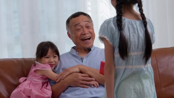 サプライズ高齢の祖父母 陽気な高齢男性を笑顔にし お祝いのための孫娘からのサプライズギフトを受け取っている間 後ろにプレゼントボックスを隠している小さな少女 — ストック動画