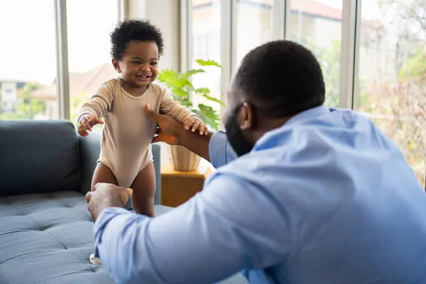 Portrait Of Happy African American Dad With Cute Little Baby Girl on couch at home in the living room, caring father smiling and amusing his girl while sitting on the couch, happy family