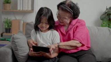 Sevinçten gülümseyen olgun bir büyükanne ve küçük torunu rahat koltukta tablet kullanıyor. Mutlu yetişkin büyükanne ve küçük torun eğitim videoları oynuyor. Çevrimiçi eğitim, olgun eğlence kavramı.