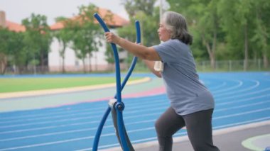 Kıdemli Asyalı kadın halka açık parktaki egzersiz istasyonlarında günlük antrenman yapıyor. Fiziksel aktivitelerle genç kalmayı, açık hava spor salonunu temsil ediyor. Fiziksel sağlık konsepti. Şehir parkında açık hava fitness ekipmanları