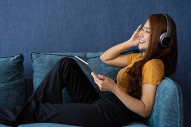 Kulaklıklı, rahat, genç Asyalı bir kadın akıllı telefonuyla online müzik medyası eğlencesi dinliyor, evdeki kanepede dinleniyor. Düz dinlenme, oturma odasında boş zaman geçir. Salon konsepti