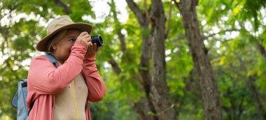 Sırtında kamera ve sırt çantası tutan Asyalı olgun bir kadının portresi. Asya 'da etkin bir son sınıf öğrencisi parkta doğanın tadını çıkarıyor. Açık havada bir ormanda bir patikada dikiliyorum. Aktif seyahatin tadını çıkarıyorum.