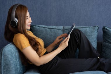 Kulaklıklı, rahat, genç Asyalı bir kadın akıllı telefonuyla online müzik medyası eğlencesi dinliyor, evdeki kanepede dinleniyor. Düz dinlenme, oturma odasında boş zaman geçir. Salon konsepti