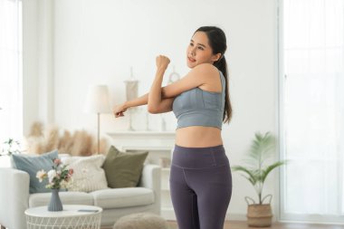 Spor giyimli genç bir Asyalı kadın sağlık, spor ve sağlık için evinde yoga yapıyor, ev içi spor eğitiminden ve sağlıklı yaşam tarzından keyif alıyor, şık giyiniyor ve gülümsüyor.