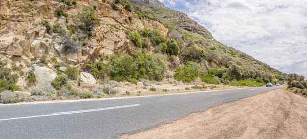 False Bay batı kıyısındaki sarkık renkli kayaların altında uzanan kıyı yolu manzarası, parlak yaz ışığında, Cape Town, Batı Cape, Güney Afrika