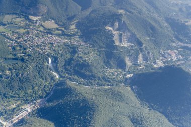 Kuzeyden Collestatte, Apennines, Terni, Umbria, İtalya yakınlarındaki parlak yaz ışığında çekilen, dağlık yamaçların arasında köy ve yüksek şelaleli bir hava manzarası.