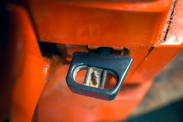 The lever of the air suction cable for cold starting the engine on the body of an orange chainsaw close-up. Rich-fuel mixture choke button for engine start