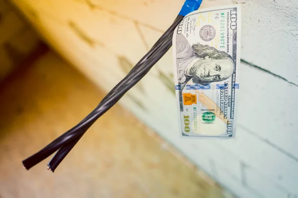 A hundred dollar bill hangs on an electrical cable attached to a bare aerated concrete brick wall close-up against a blurred background. Payment for electrical services. Rising prices for electricity
