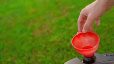 Çim biçme makinesinin deposundaki yakıt deposu yeşil bir çimenliğin arka planına karşı. Bir el, çim biçme makinesini benzinle doldurduktan sonra kırmızı plastik bir huniyi yakına çeker. Yüksek kalite FullHD