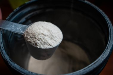 Toz kavanozunun arka planında beyaz tozla dolu bir kepçeyi tutan el. Spor beslenme girişi.