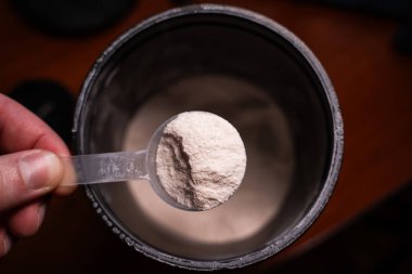 Toz kavanozunun arka planında beyaz tozla dolu bir kepçeyi tutan el. Spor beslenme girişi.