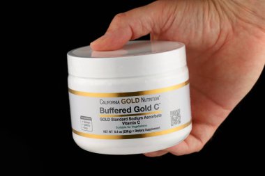 LVIV, UKRAINE - May 22, 2024: Hand holding Buffered Gold C supplement container by California Gold Nutrition against black background. clipart