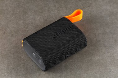 LVIV, UKRAINE - September 09, 2024: A compact portable speaker rests on a gray surface, showcasing its sleek design and textured fabric. The vibrant orange strap adds a pop of color to the product. clipart