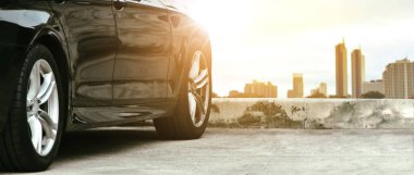 Şehrin arka planındaki siyah arabayı güneş ışığıyla kapat. Otoparka park etmiş araba, panoramik pankart, ulaşım konsepti.