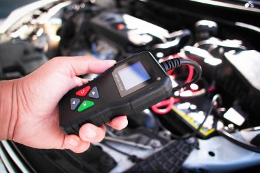 Araba Aküsü Teşhis Aracı, bir teknisyen araç aküsü testçisini açık bir arabanın kaputunun üzerinde tutuyor, motor görünür durumda, aracın aküsünü kontrol etmek için..