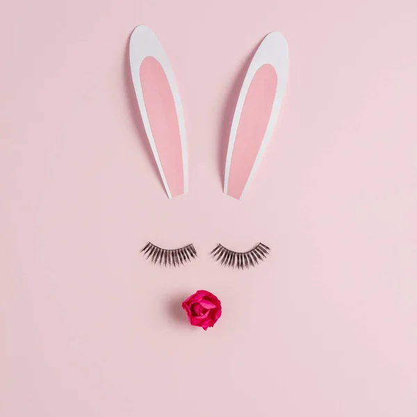 Easter bunny face made of rabbit ears with eyelashes and rose flower on pink background. Happy Easter minimal concept.