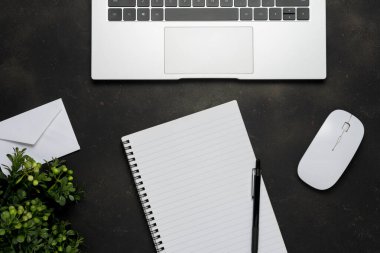 Çalışma alanı boş defter, dizüstü bilgisayar, ofis malzemeleri ve siyah arkaplanda yeşil yapraklar.