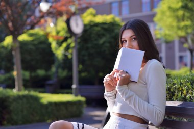 Güzel kahverengi gözlü, beyaz, boş kaplı bir kitap tutan, kameraya bakarken dudaklarını saklayan güzel bir kadının portresi. Şehir parkında ağaçlar ve bitkilerle çevrili bir bankta oturuyor.