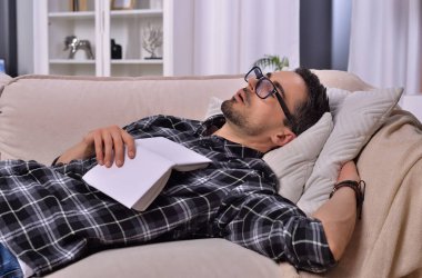 Kitaplık sükuneti. Yüzünde gözlük olan genç bir adam oturma odasındaki lüks bir kanepede kitap okurken uyuyakaldı. Rahatlama ve edebiyat kavramı.