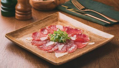 İtalyan Biftek Carpaccio, ahşap bir masada roka ile süslenmiş, kare bir tabakta.