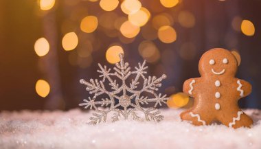 A gingerbread man and a sparkly silver snowflake sit on a snowy surface against a blurred background of twinkling lights clipart