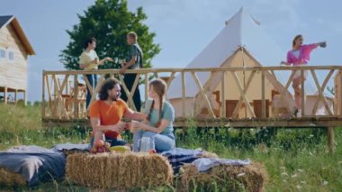 Kameranın önünde kameraya poz veren harika görünümlü bir çift arka plandaki samanlıkta otururken diğer arkadaş grupları çadırın yanında eğleniyor. 4k