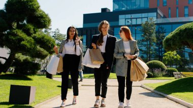 Karizmatik güzel bayan arkadaşlarıyla caddenin ortasında buluşuyor. Üç bayan kameranın önünde yürümekten ve elinde ekolojik alışveriş torbalarıyla konuşup gülümsüyor.