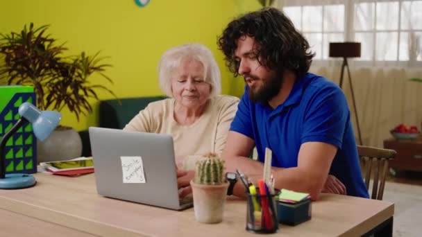 若い男はノートパソコンを使用して彼女の息子と一緒にリビングルームの幸せな老婦人の家では 古い女性にノートパソコンを使用する方法を説明します — ストック動画