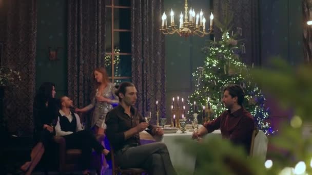两个英俊俊俏的男人和朋友一起庆祝圣诞节或新年 他们坐在圣诞餐桌旁聊天 而其他的朋友则坐在背景上聊天 — 图库视频影像