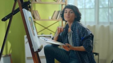 Mavi flanel tişört giyen güzel bir kadın büyük ve parlak bir sanat atölyesinde tuvaline inanılmaz ve modern bir şey çiziyor. ARRI Alexa Mini 'de çekilen