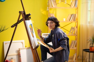 Mavi flanel tişört giyen güzel bir kadın büyük ve parlak bir sanat atölyesinde tuvaline inanılmaz ve modern bir şey çiziyor. Portre