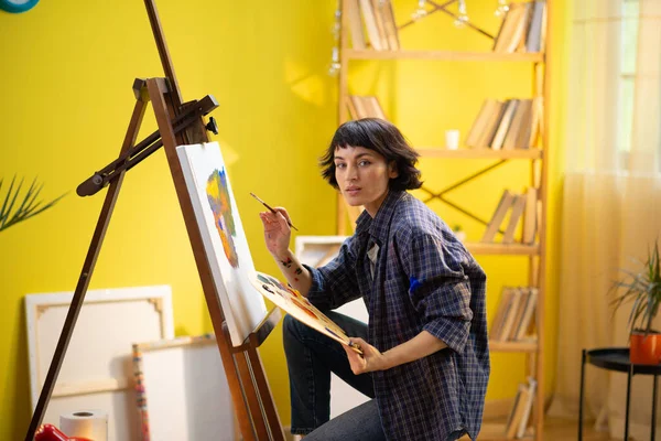 Mavi flanel tişört giyen güzel bir kadın büyük ve parlak bir sanat atölyesinde tuvaline inanılmaz ve modern bir şey çiziyor. Portre