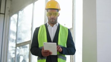 Good looking young architect man with safety equipment at construction site using digital tablet to analysing the plan of construction.