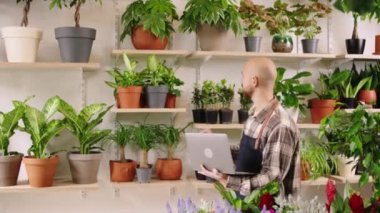 In the floral store the florist man the owner of the shop walking through the flowers shop and checking with the laptop the inventory of the shop he is the owner of shop. 4k