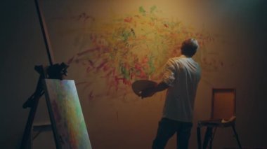 Talented artist man in his art studio painting modern artwork on the wall with fingers he holding the colour palette on hand. 4k