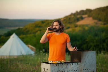 Bir adam takla atmaya ve farklı sebzeleri doğadaki ızgaraya pişirmeye odaklanırken çok eğleniyor. Portre