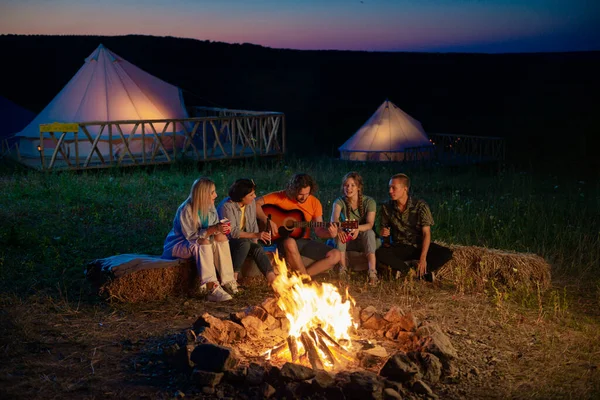In the night time at the campsite group of multiracial friends sitting down on the haystack playing on the guitar singing and dancing feeling relaxed and excited. Tent