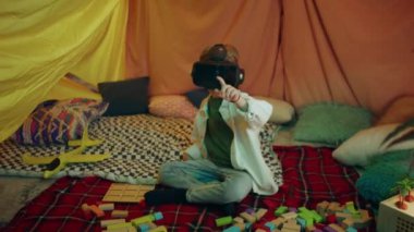 A young cute boy looks very happy as he is playing intensely with a vr headset inside his cosy looking indoor sheet tent.