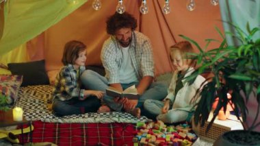 Two cute boys are having fun with their dad as they are bonding together and reading a book in an indoor sheet tent. 4k