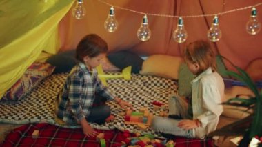 Two very adorable young boys are playing with building blocks inside a huge blanket tent.