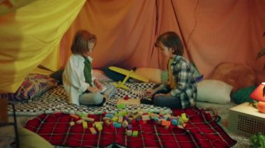 In a very comfy and cozy looking indoor blanket tent filled with pillows there are two boys playing with colorful building blocks.