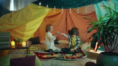 Two little boys who get along very well are playing around with a virtual reality headset in their own huge colourful blanket tent.