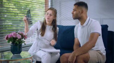 Charismatic woman doctor have a conversation with her patient man they discussing about his results on the x-ray while sitting down on the sofa. 4k