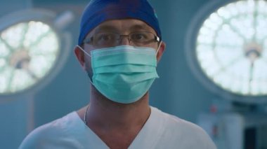 A close up shot of a charming male surgeon wearing glasses and a blue medical face mask.