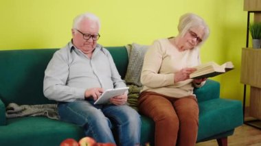 Perfect looking old couple speeding time at home sitting on the sofa in living room man using the digital tablet to watch something and beautiful woman read a book. Shot on ARRI Alexa Mini
