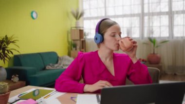 In front of the camera at home charismatic woman working online on her laptop while she have break she take her headphones listen music dancing and drinking some coffee. 4k