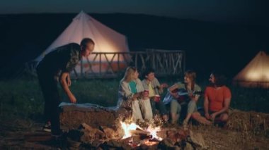 In front of the camera at the nature at campsite group of ladies and guys very handsome and attractive sitting down on the haystack playing on the guitar and drinking something beside the fire stake